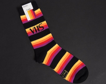 May include: Black socks with horizontal stripes in shades of purple, orange, and yellow. The word "VHS" is printed in purple on the sock. A tag at the top has the text "FACTORY" and "F-025".