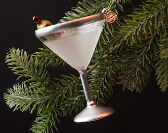 Op de afbeelding: Een zilveren martini-glas ornament met een groene olijf en een rode kers op een steel. Het ornament hangt aan een groene dennentak.