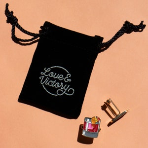 May include: Gold cufflinks with a black velvet pouch. The cufflinks feature a colorful illustration of a cocktail drink with a cherry and orange slice. The pouch has the text "Love & Victory" printed on it.