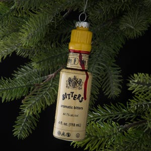 May include: A Christmas tree ornament shaped like a bitters bottle. The ornament has a yellow cap, a label with the word "Bitters", and a red stripe. The bottle is 4 fl. oz. (118 mL).