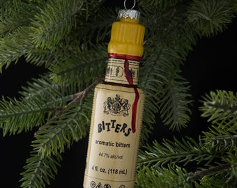 Op de afbeelding: Kerstboomversiering in de vorm van een bittersfles. De versiering heeft een gele dop, een etiket met het woord "Bitters" en een rode streep. De fles is 118 ml.
