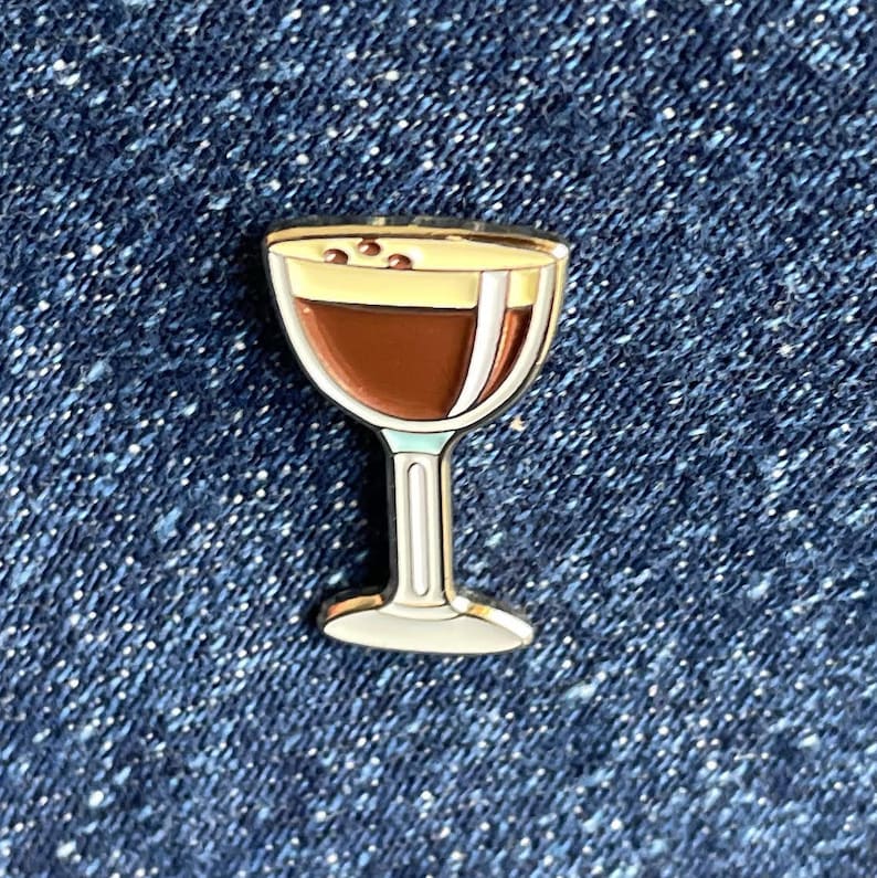 Op de afbeelding: Een emaille pin in de vorm van een cocktailglas. Het glas is gevuld met een donkerbruine vloeistof, bedekt met een lichtgele schuimlaag en drie kleine bruine stippen. De pin heeft een witte steel en voet, tegen een blauwe denim achtergrond.