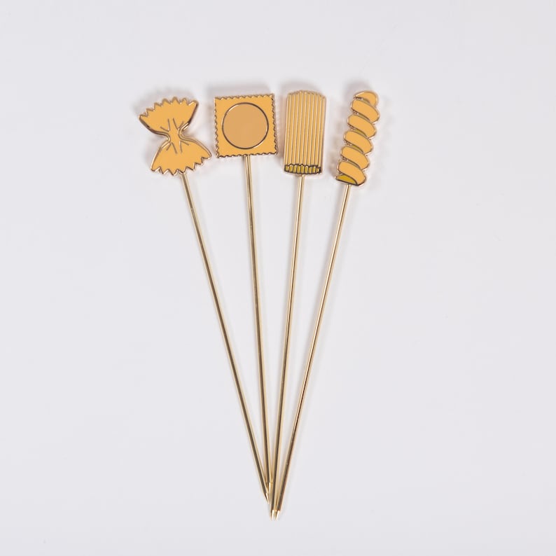 May include: Four pasta-shaped appetizer picks with gold stems and yellow pasta designs. The pasta shapes include farfalle, a square ravioli, a ridged pasta, and a spiral pasta. The picks are arranged on a white background.