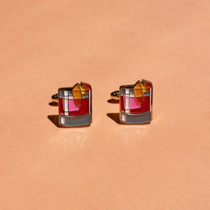 May include: A pair of gold cufflinks with a red, pink, and orange cocktail drink design.