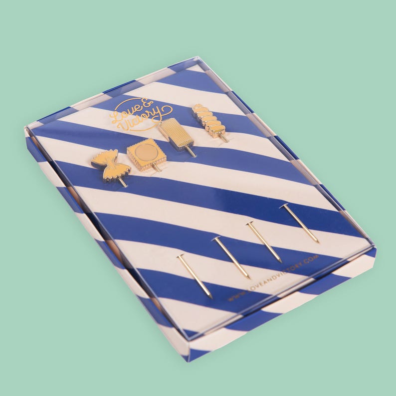 May include: A set of gold-colored pins in a clear box with a blue and white striped pattern. The pins include various shapes, such as a bow tie, a square, and a popsicle. The box has the text "Love & Victory" in gold.