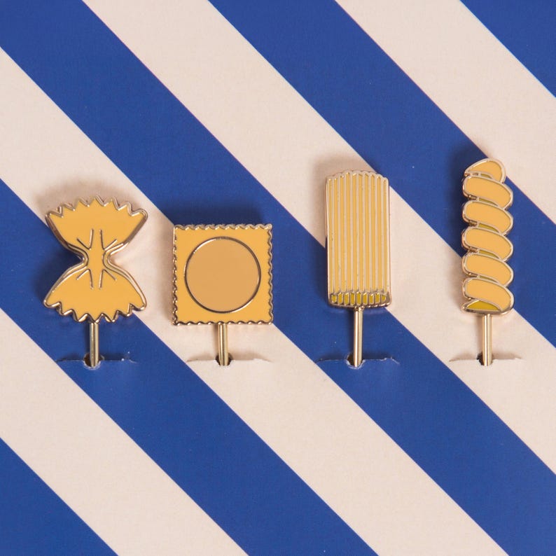 May include: Four yellow pasta-shaped pins on a white and blue striped background. The pins include a farfalle, a square with a circle, a rectangular shape, and a spiral shape. Each pin has a silver pin backing.