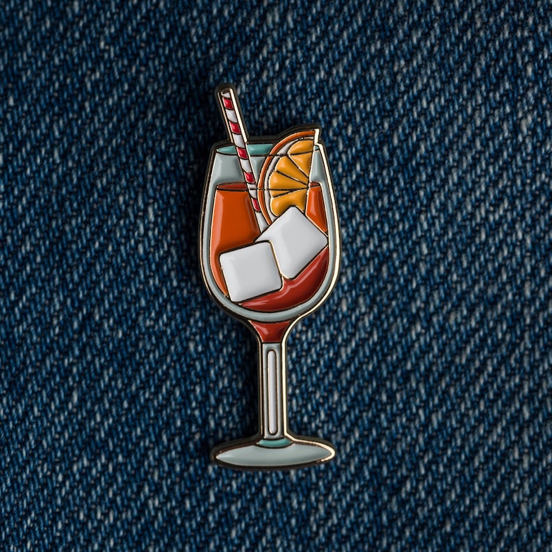 May include: A gold-coloured enamel pin of a glass of orange juice with ice cubes and a red and white striped straw. The glass is filled with a bright orange liquid and has a gold rim.