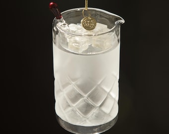 Op de afbeelding: Een heldere glazen cocktailornament met een ruitpatroon, gevuld met ijs en vloeistof. Een kleine gouden bedel hangt in het glas. Een rode roerstaaf ligt op de rand. Het ornament hangt tegen een zwarte achtergrond.