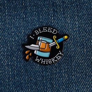 May include: A black enamel pin with a cartoon illustration of a glass of whiskey with a dagger through it. The text "I Bleed Whiskey" is written around the glass.