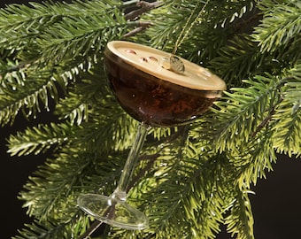 Op de afbeelding: Een decoratieve kerstversiering in de vorm van een cocktailglas. Het glas is bruin met een beige bovenkant en hangt aan een gouden draad. De versiering staat tegen een achtergrond van groene dennennaalden.