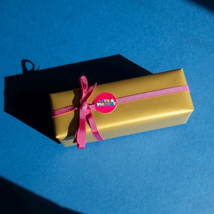 Pu&ograve; includere: Un cofanetto regalo dorato, legato con un nastro rosa e un adesivo abbinato con la scritta "LOVE & VICTORY" su sfondo blu. Il cofanetto regalo &egrave; rettangolare e proietta un'ombra sulla superficie blu.