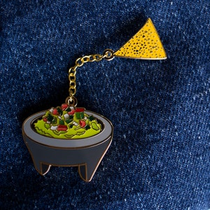Può includere: Uno spillo in metallo dorato con un guacamole verde, rosso e bianco in una ciotola grigia e un chip triangolare giallo.