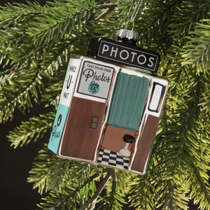 Può includere: Un ornamento natalizio a forma di cabina fotografica con la scritta "PHOTOS" in alto. L'ornamento ha una combinazione di colori turchese, marrone e bianco, con il testo "TAKE YOUR OWN PHOTOS Only $5". L'ornamento è appeso a un ramo di pino verde.