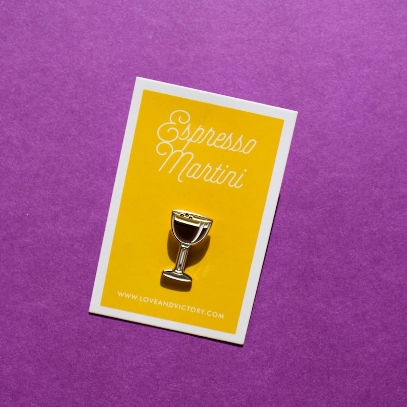 Op de afbeelding: Een zilverkleurige reversspeld in de vorm van een espresso martiniglas, geplaatst op een gele kaart met de woorden "Espresso Martini" in cursief. De kaart staat op een paarse achtergrond. Het websiteadres www.loveandvictory.com staat onderaan de kaart.