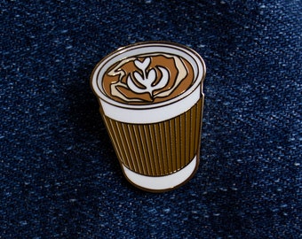 Peut inclure: Une épingle en émail dorée représentant une tasse à café blanche avec un manchon en papier brun. Le café a un motif de latte art brun et blanc.