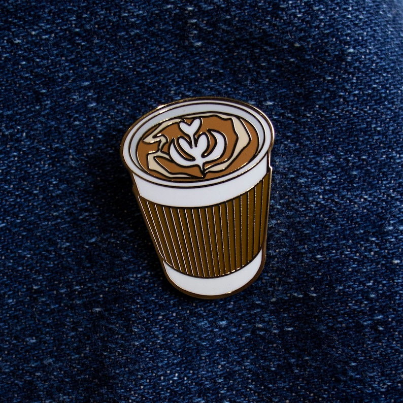 May include: A gold-colored enamel pin of a white coffee cup with a brown paper sleeve. The coffee has a brown and white latte art design.
