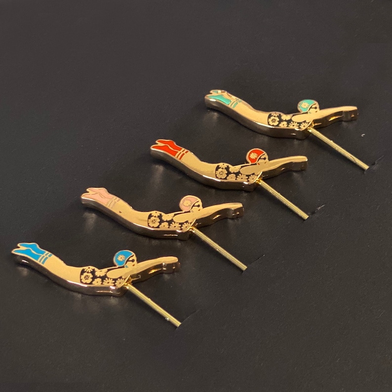 May include: Four gold-coloured metal pins featuring a black and white design of a swimmer diving into water. Each pin has a different coloured accent on the swimmer's cap and legs: blue, pink, red, and green.