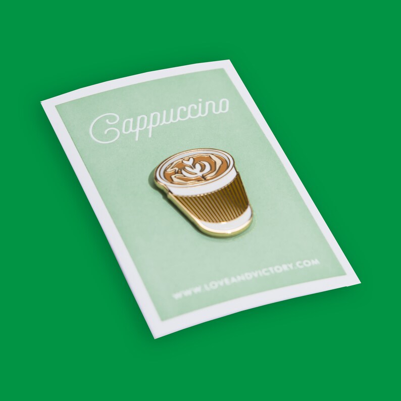 May include: Gold-tone enamel pin featuring a white and brown cappuccino cup with latte art. The pin says "Cappuccino" and "www.loveandvictory.com".