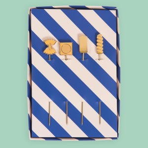 May include: A set of four gold-colored appetizer picks in a blue and white striped box. Each pick features a different food-themed design: bow tie pasta, a stamp, a popsicle, and a spiral. The box is rectangular and has a light blue background.