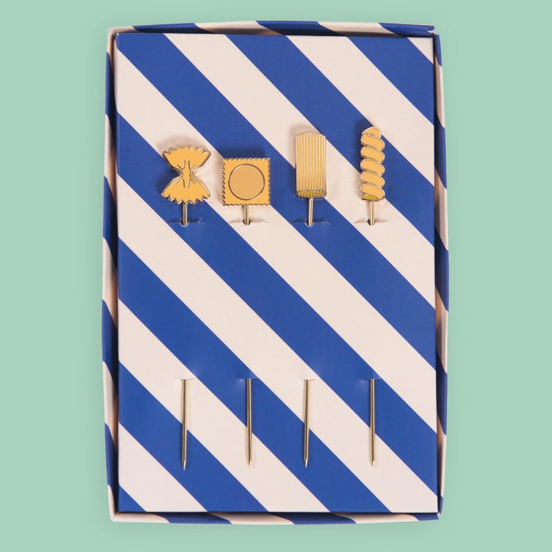 May include: A set of four gold-colored appetizer picks in a blue and white striped box. Each pick features a different food-themed design: bow tie pasta, a stamp, a popsicle, and a spiral. The box is rectangular and has a light blue background.