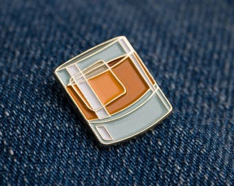 May include: A gold-colored enamel pin shaped like a glass of whiskey. The glass is light blue with a white outline and the whiskey is orange.