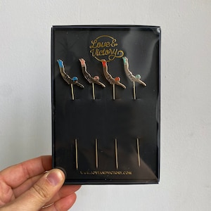May include: Set of five gold-toned pins featuring a diver in different colours. The pins are packaged in a black box with the text "Love & Victory" and "www.loveandvictory.com".