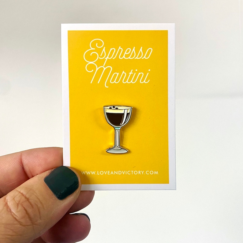 Op de afbeelding: Een emaille pin met een espresso martini ontwerp. De pin staat op een gele kaart met de woorden "Espresso Martini" in cursief. De pin is een cocktailglas met een donkerbruin drankje en koffiebonen. Het websiteadres is zichtbaar.