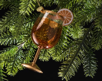Op de afbeelding: Een gouden glazen ornament in de vorm van een cocktailglas met een schijfje sinaasappel aan de rand. De ornament hangt aan een groene kunstmatige dennentak.