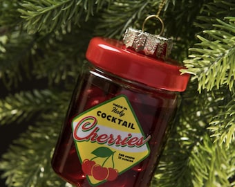 Op de afbeelding: Glazen kerstbal in de vorm van een potje gevuld met rode vloeistof en kersen, met een rode deksel en een goudkleurige metalen dop. Het etiket luidt "Made in Italy Cocktail Cherries". De ornament bevat 340 gram of 12 ounces.