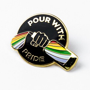 May include: Gold-colored enamel pin with a black background. The pin features a fist holding a bottle with a rainbow design. The text "POUR WITH PRIDE" is written around the edge of the pin.