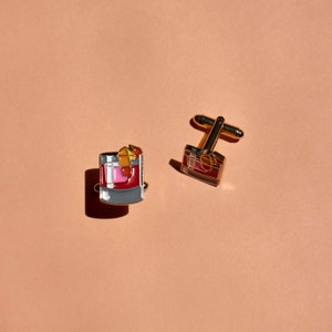 May include: A pair of gold cufflinks. One cufflink features a red, pink, and yellow cocktail drink. The other cufflink is a simple gold rectangle.