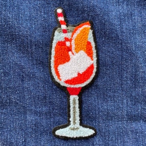 Puede incluir: Una aplicación de fieltro de una bebida naranja con una pajita roja y blanca y una rodaja de naranja. La bebida está en un vaso con un tallo y una base redonda.