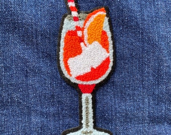 May include: A felt applique of an orange drink with a red and white straw and an orange slice. The drink is in a glass with a stem and a round base.