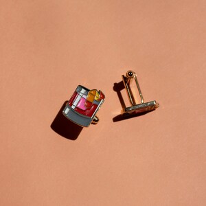 May include: A pair of gold cufflinks. One cufflink features a colorful illustration of a cocktail drink. The other cufflink is a simple gold design.