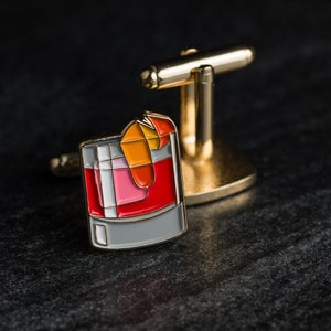 May include: Gold cufflinks with a red, orange, and pink cocktail drink design. The drink is in a glass with a gray rim.