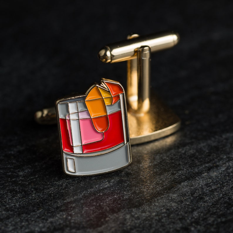 May include: Gold cufflinks with a red, orange, and pink cocktail drink design. The drink is in a glass with a gray rim.