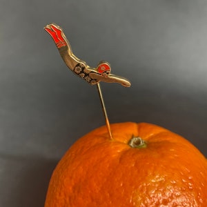 May include: A gold-coloured pin of a diver in a red swimsuit and a black and white floral pattern. The diver is wearing a red and white swim cap. The pin is stuck into an orange.