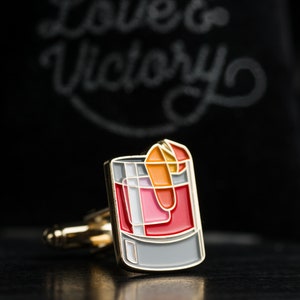 May include: Gold cufflinks with a red, orange, and white enamel design of a cocktail glass. The cufflinks are on a black surface with the text "Love & Victory" in white chalk.