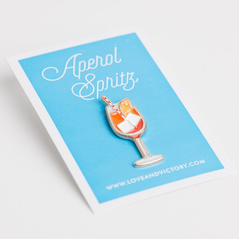 May include: A blue card with white text that reads "Aperol Spritz" and a gold enamel pin of an orange drink with a straw and ice cubes. The website www.loveandvictory.com is printed on the card.