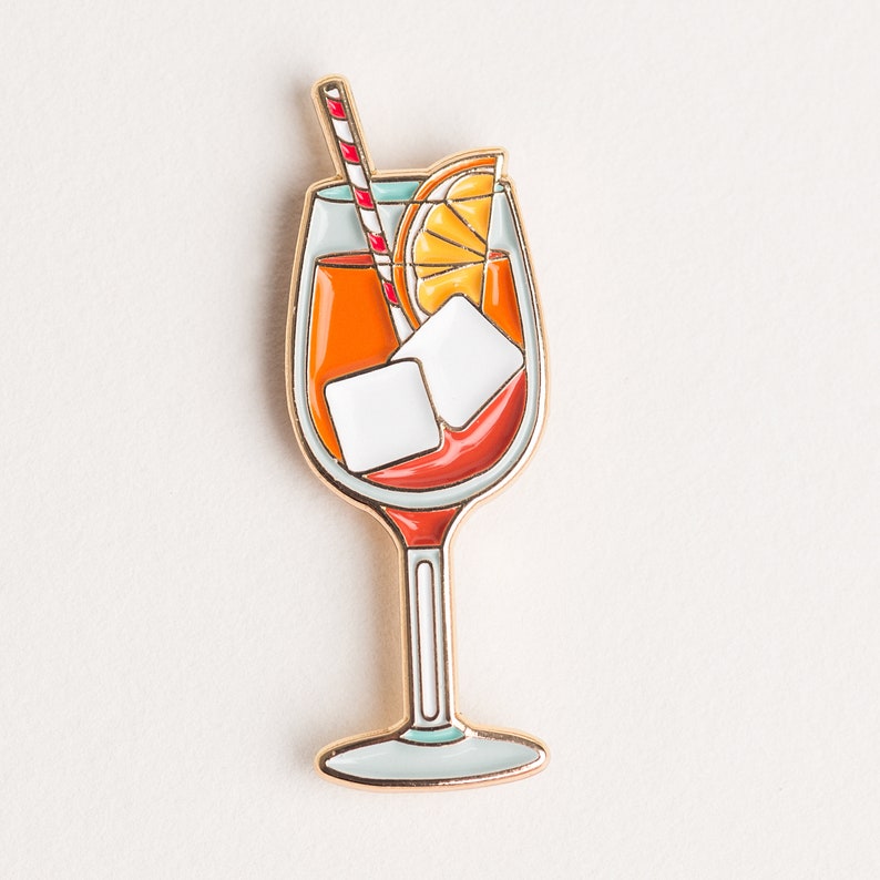 May include: Gold-toned enamel pin of a glass of Aperol Spritz with an orange slice, ice cubes, and a red and white striped straw.