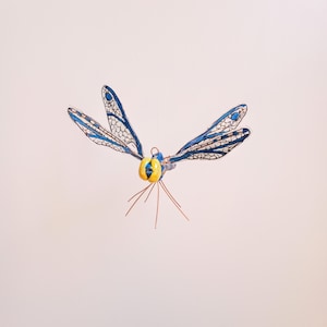 May include: A blue and white dragonfly with yellow eyes, made of wire and glass, is suspended in mid-air against a white background.
