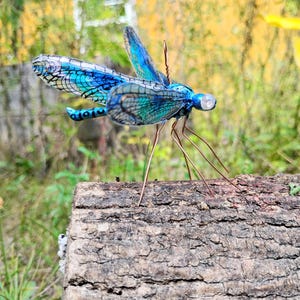 Puede incluir: Una escultura artesanal de libélula con alas iridiscentes azules y turquesas. La libélula tiene patas y antenas de alambre, y está posada sobre madera envejecida. Las alas tienen un patrón detallado y veteado. La escultura mide unos 15 cm de largo.