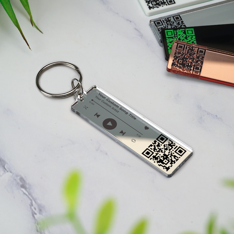 May include: Silver keychain with a clear acrylic charm featuring a music player design. The charm has a play button, rewind and fast forward buttons, and a heart icon. It also has a QR code and the text "Your Custom Song Title" and "Your Custom Artist".