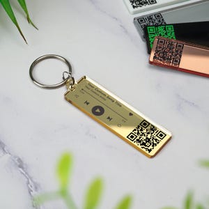 May include: Gold-colored keychain with a custom song title and artist, a play button, and a QR code. The keychain is attached to a silver key ring.