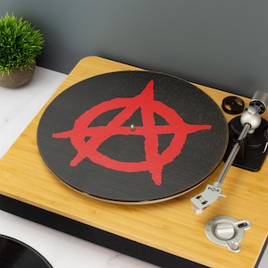 Może przedstawiać: Czarny gramofon z czerwonym symbolem anarchii na talerzu. Gramofon jest wykonany z drewna i ma srebrny tonearm.