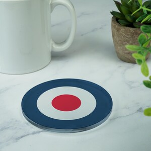 Op de afbeelding: Een ronde glazen onderzetter met een blauw, wit en rood doelwitontwerp. De onderzetter ligt op een wit marmeren oppervlak.
