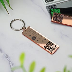May include: Rose gold keychain with a custom song title, artist, and a QR code. The keychain has a play button, rewind button, and fast forward button.