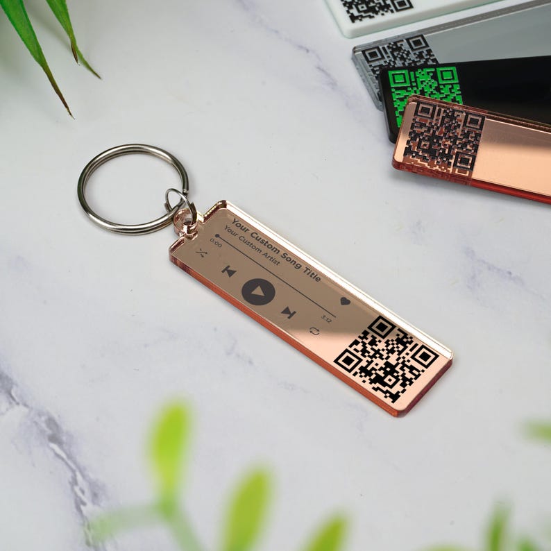 May include: Rose gold keychain with a custom song title, artist, and a QR code. The keychain has a play button, rewind button, and fast forward button.