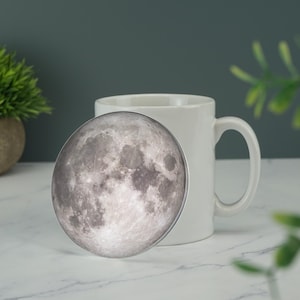 Peut inclure: Une tasse en céramique blanche avec une image couleur de la lune sur le devant. L'image de la lune est nette et la tasse est légèrement floue.