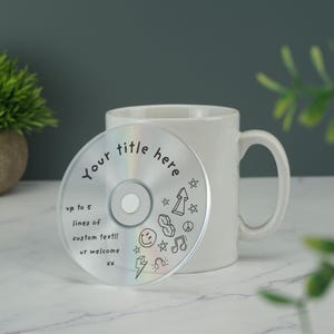 May include: A white ceramic mug with a blank space for text and a CD on top of the mug. The CD has a rainbow iridescent surface and black line drawings of stars, a peace sign, a smiley face, musical notes, an arrow, and a lightning bolt. The text on the CD reads "Your title here" and "up to 5 lines of custom text!! ur welcome XX".